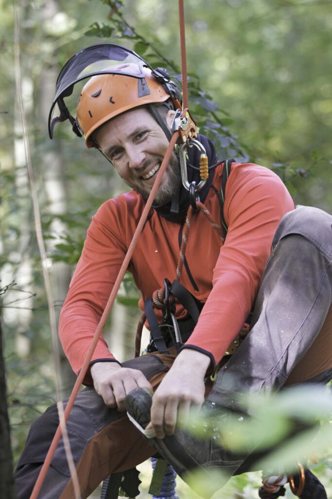 Kontakt freudiger Baumpfleger hängt mit professioneller Ausrüstung im Kletterseil